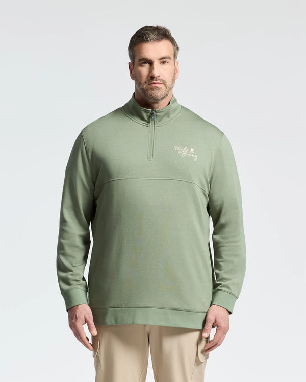 A man wearing a Psycho Bunny desert sage green 1/4 mock zip pullover stands against a white background. The cotton jersey top pairs well with his light khaki pants. He has short hair and a beard, completing the polished look.