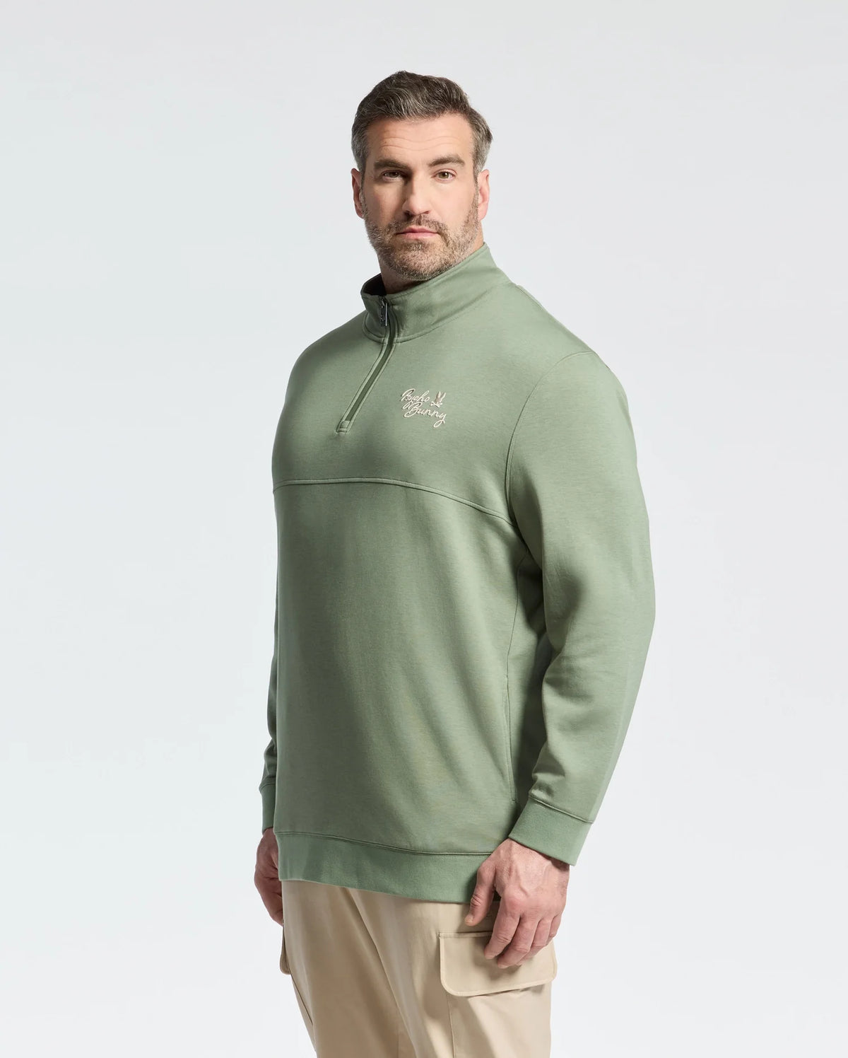 A man with short hair and a beard wears a Psycho Bunny Men's Big and Tall Desert 1/4 Mock Zip sweatshirt in light green with an embroidered wordmark logo on the chest. He pairs it with beige cargo pants, standing against a plain white background.