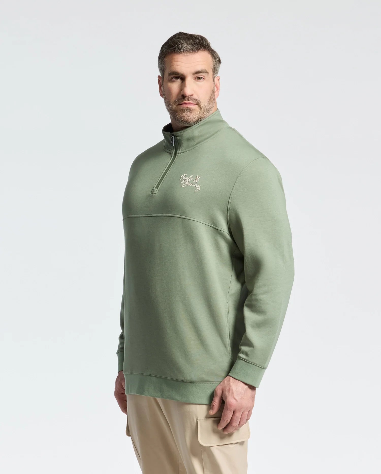 A man with short hair and a beard wears a Psycho Bunny Men's Big and Tall Desert 1/4 Mock Zip sweatshirt in light green with an embroidered wordmark logo on the chest. He pairs it with beige cargo pants, standing against a plain white background.
