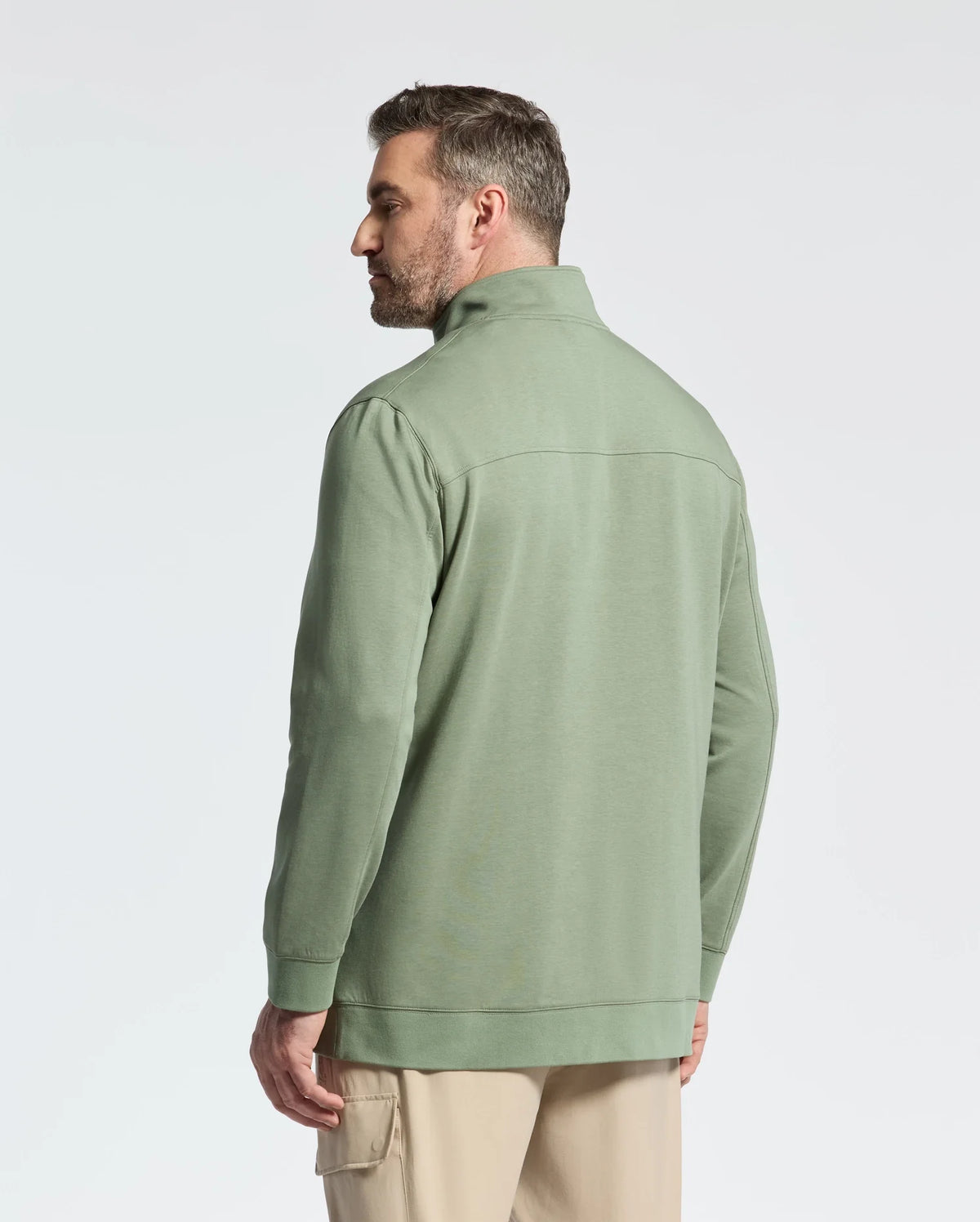 A man with short dark hair, in profile facing left, wears a sage green Psycho Bunny shirt featuring a wordmark logo and beige cotton jersey pants. The plain background is light-colored.