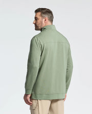 A man with short dark hair, in profile facing left, wears a sage green Psycho Bunny shirt featuring a wordmark logo and beige cotton jersey pants. The plain background is light-colored.