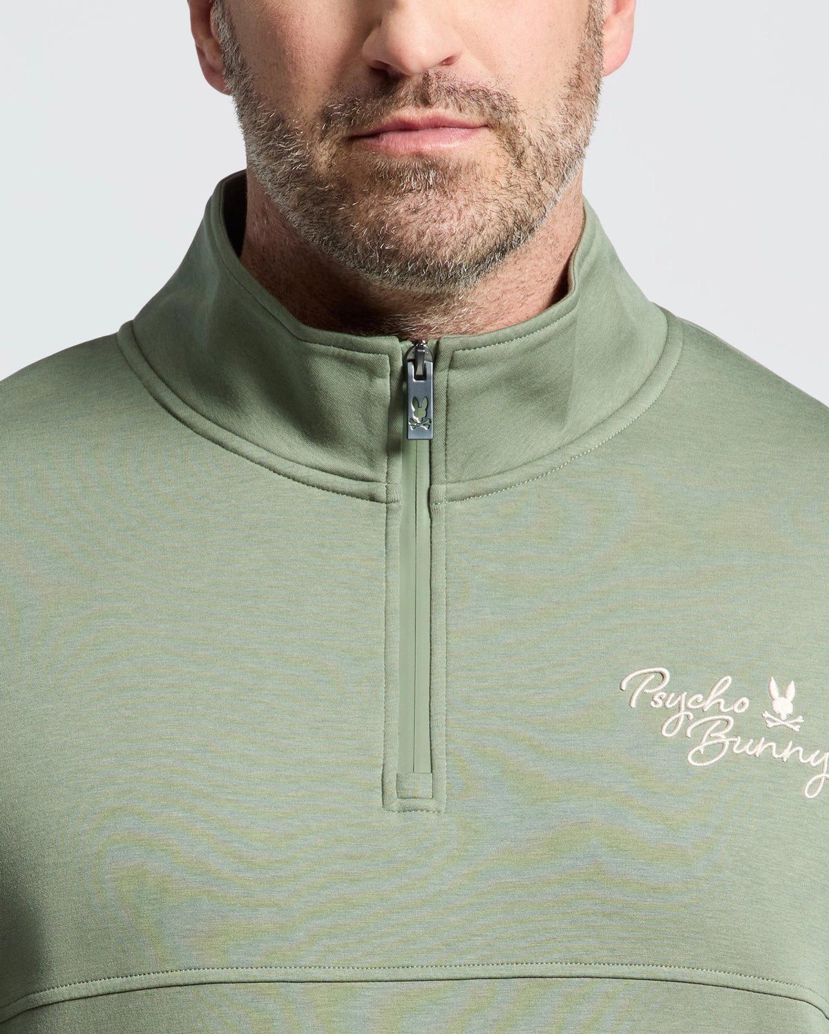 A man wears the Psycho Bunny Men's Big and Tall Desert 1/4 Mock Zip (B9S488F200) in sage green, featuring a logo on the chest. Made from soft cotton jersey, it has a standing collar and quarter-zip closure, set against a neutral grey backdrop.