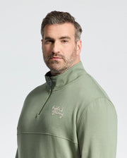 A man in a sage green Psycho Bunny cotton jersey quarter-zip sweatshirt with an embroidered "Golf Bunny" wordmark stands against a plain background. He has short hair and facial hair, looking directly at the camera.
