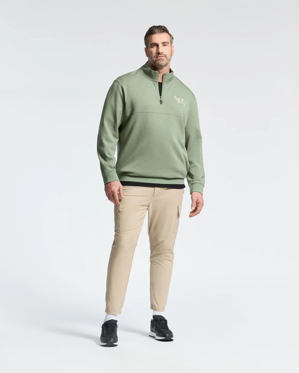 A man in the Psycho Bunny Men's Big and Tall Desert 1/4 Mock Zip stands against a plain background, paired with beige pants, black shoes, and white socks. His hands rest by his sides as he gazes slightly to the left. The green cotton jersey sweater features an embroidered wordmark logo.