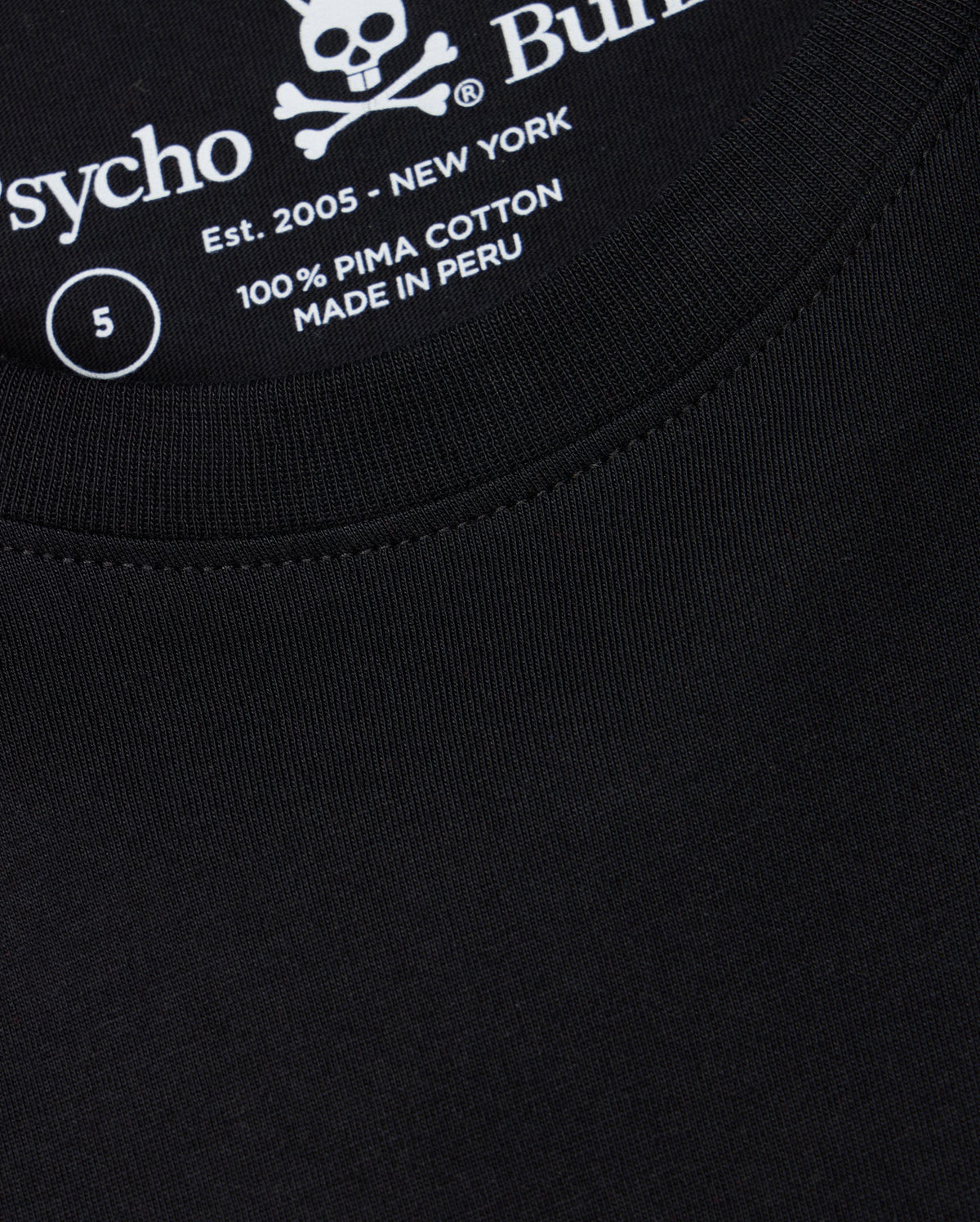 A close-up of the Psycho Bunny men's Big and Tall Classic Crew Neck Tee (B9U014CRPC) reveals a black T-shirt with white text inside featuring a skull and crossbones, the word "Psycho," "100% Pima Cotton," "Made in Peru," "Est. 2005 - New York," and size label "5.