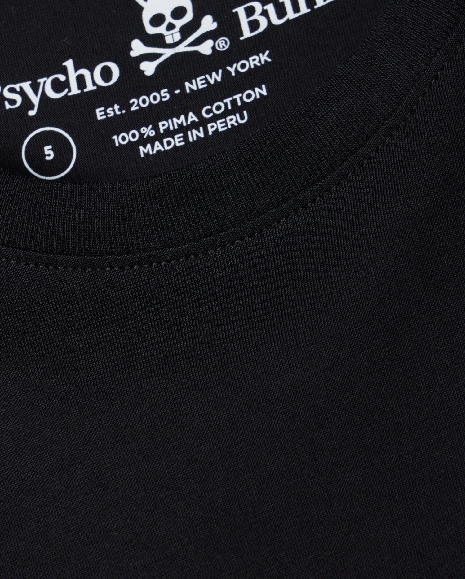 A close-up of the Psycho Bunny men's Big and Tall Classic Crew Neck Tee (B9U014CRPC) reveals a black T-shirt with white text inside featuring a skull and crossbones, the word "Psycho," "100% Pima Cotton," "Made in Peru," "Est. 2005 - New York," and size label "5.