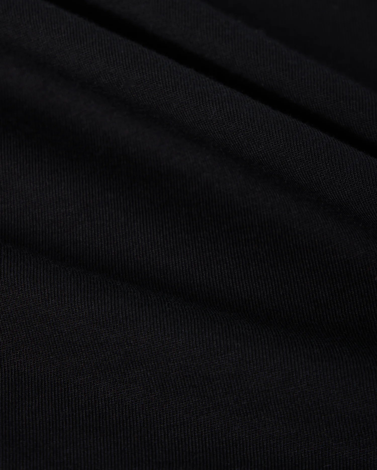 A close-up of black, finely ribbed fabric with subtle folds from the Psycho Bunny MENS BIG AND TALL CLASSIC CREW NECK TEE (B9U014CRPC) reveals a textured appearance. Lighting highlights its ridges and grooves, indicating it is soft and flexible 100% Pima cotton.
