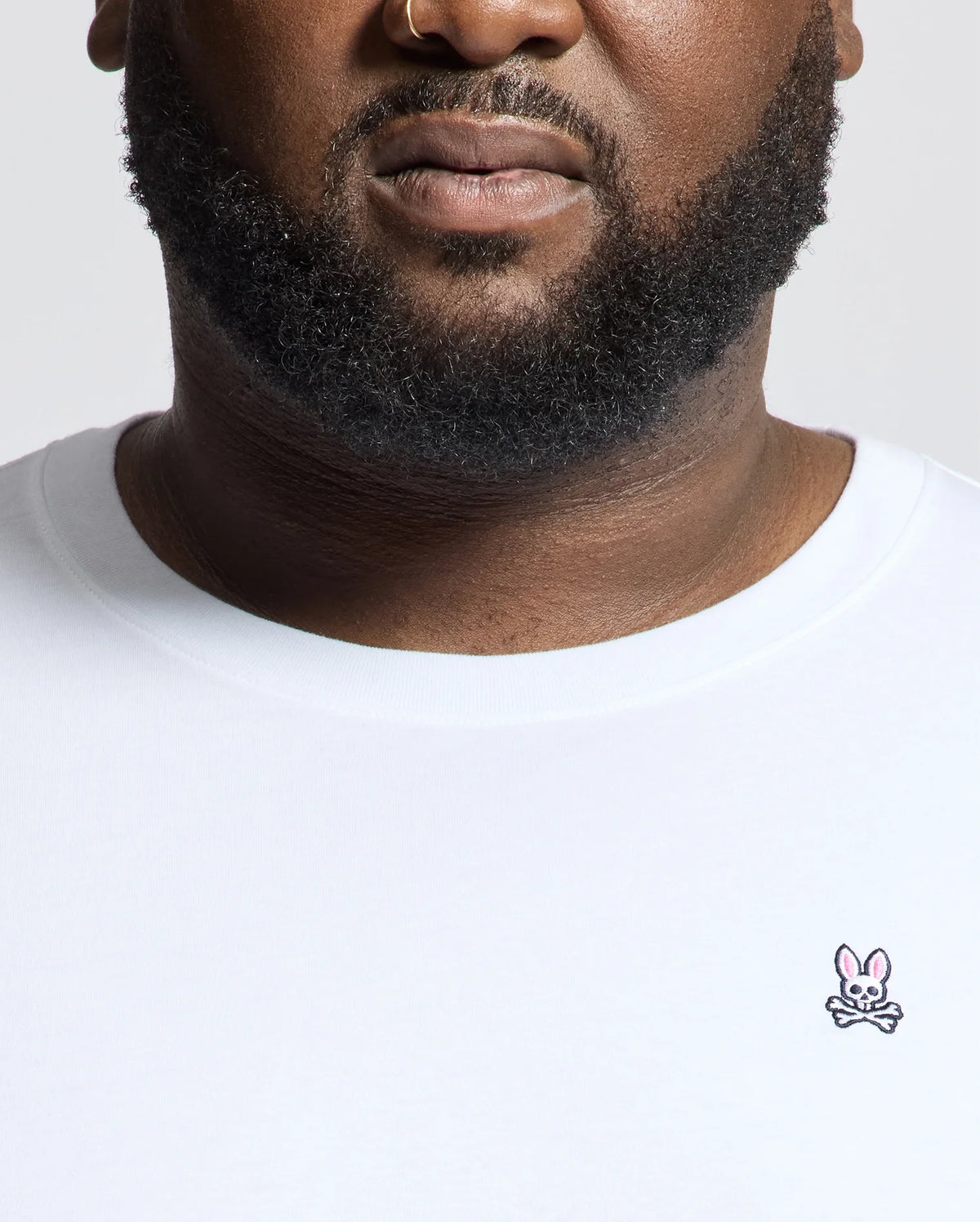 A bearded individual with a nose ring wears the Psycho Bunny MENS BIG AND TALL CLASSIC CREW NECK TEE - B9U014CRPC. It's a white Pima cotton t-shirt with a small pink bunny logo on the chest, set against a neutral backdrop—a timeless addition to any men's wardrobe.