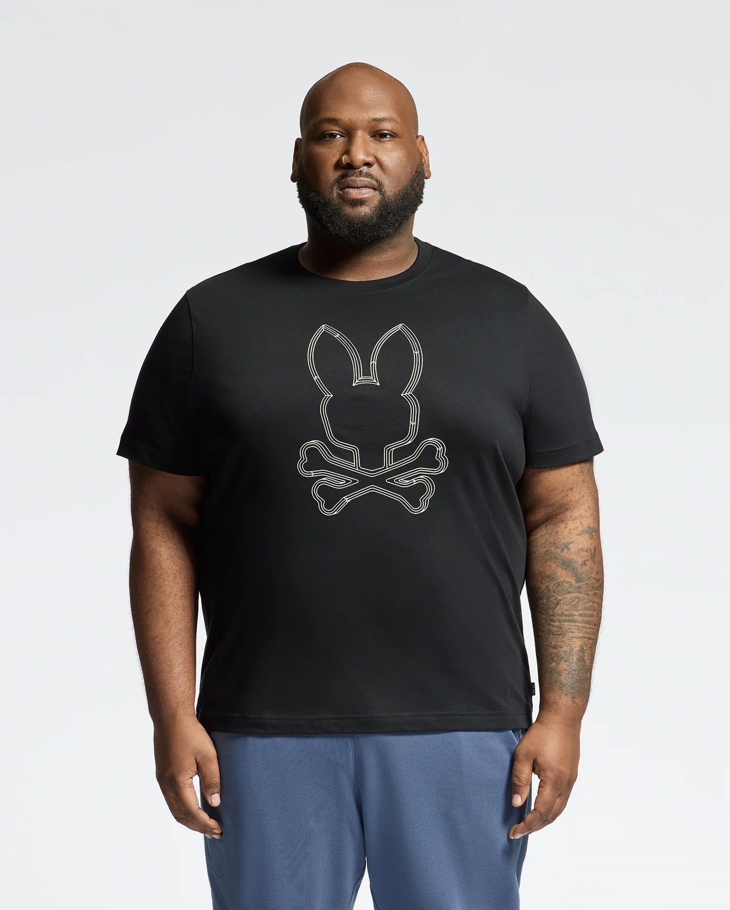 A bearded man stands against a plain backdrop, donned in Psycho Bunny's Mens Big and Tall Monarc Front Bunny Graphic Tee - B9U211E200 with its signature rabbit skull and crossbones design, paired with blue pants. His right arm is tattooed, and his expression is neutral.