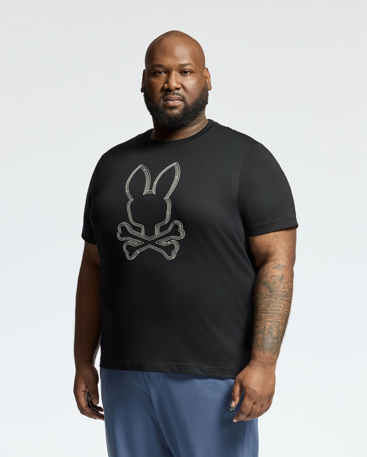 A bearded man dons a Psycho Bunny black tee, MENS BIG AND TALL MONARC FRONT BUNNY GRAPHIC TEE - B9U211E200, made from soft Pima cotton with a white rabbit and crossbones design. He reveals a tattoo on his right forearm against a plain, light-colored backdrop.