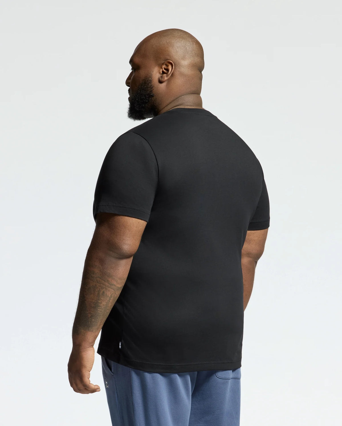 A man with a shaved head and beard, facing away from the camera, is wearing a MENS BIG AND TALL MONARC FRONT BUNNY GRAPHIC TEE by Psycho Bunny made of soft Pima cotton, paired with blue pants against a plain white background.