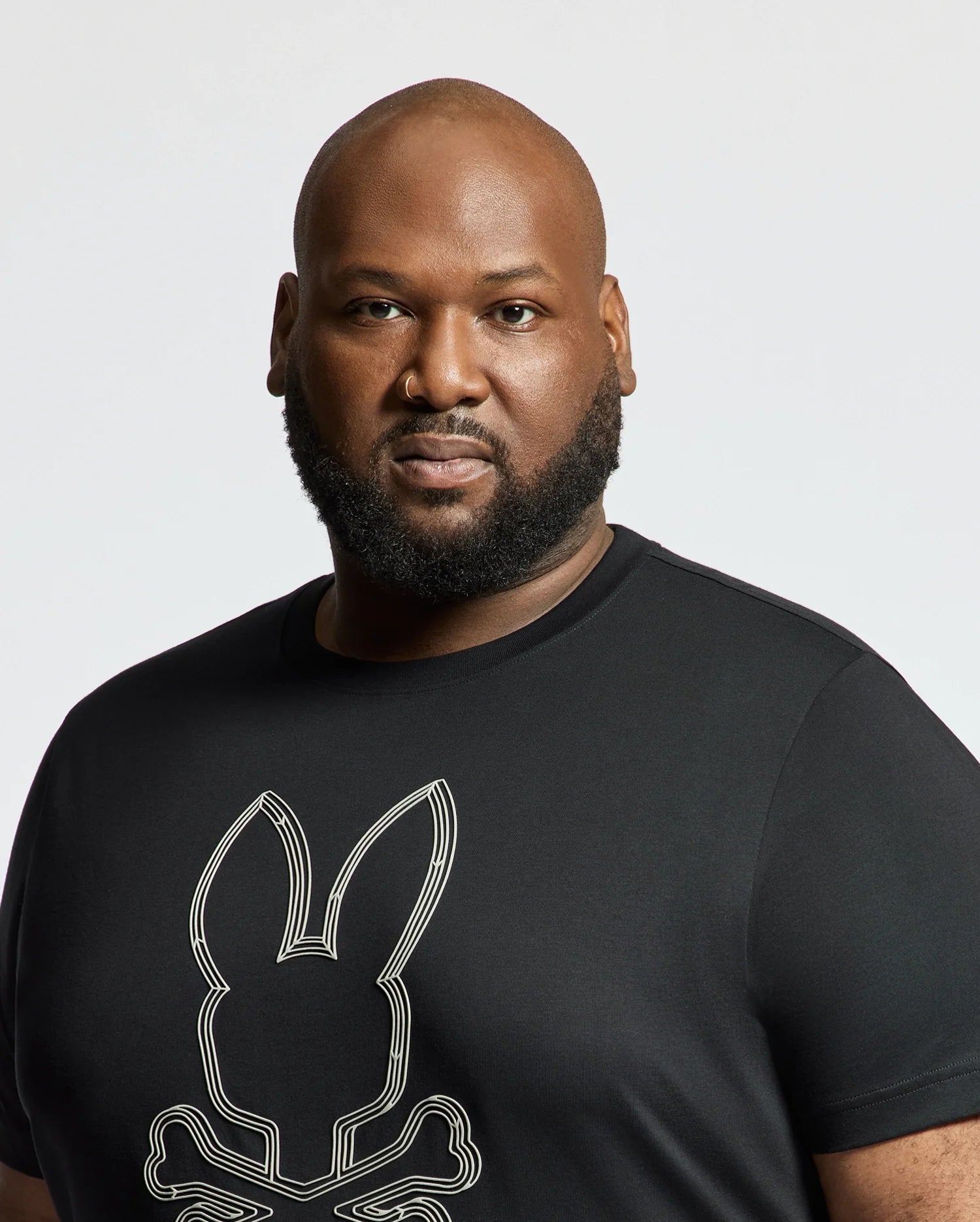 A bearded man stands against a plain white background wearing a black MENS BIG AND TALL MONARC FRONT BUNNY GRAPHIC TEE by Psycho Bunny, made of soft Pima cotton with a stylized bunny design.