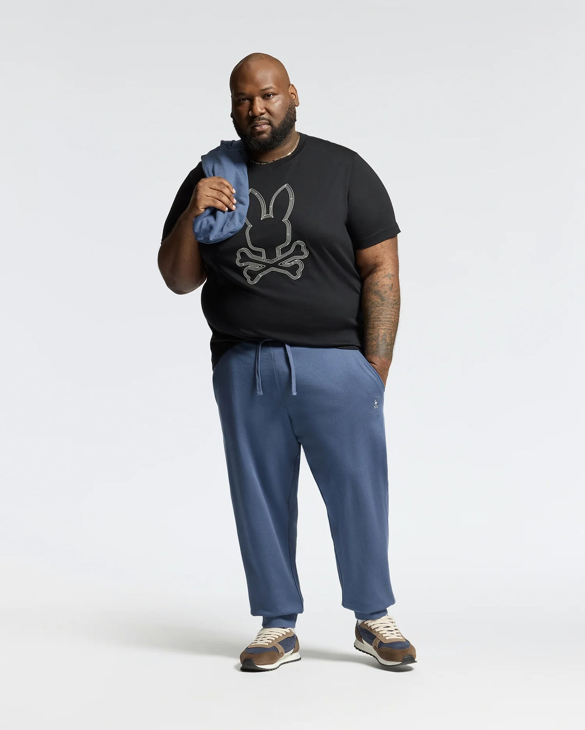A bearded man stands against a plain backdrop in a black Psycho Bunny Monarc Front Bunny Graphic Tee, paired with blue pants and brown sneakers, casually holding folded blue fabric over his shoulder.