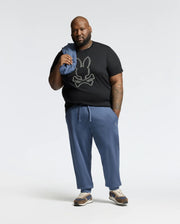 A bearded man stands against a plain backdrop in a black Psycho Bunny Monarc Front Bunny Graphic Tee, paired with blue pants and brown sneakers, casually holding folded blue fabric over his shoulder.