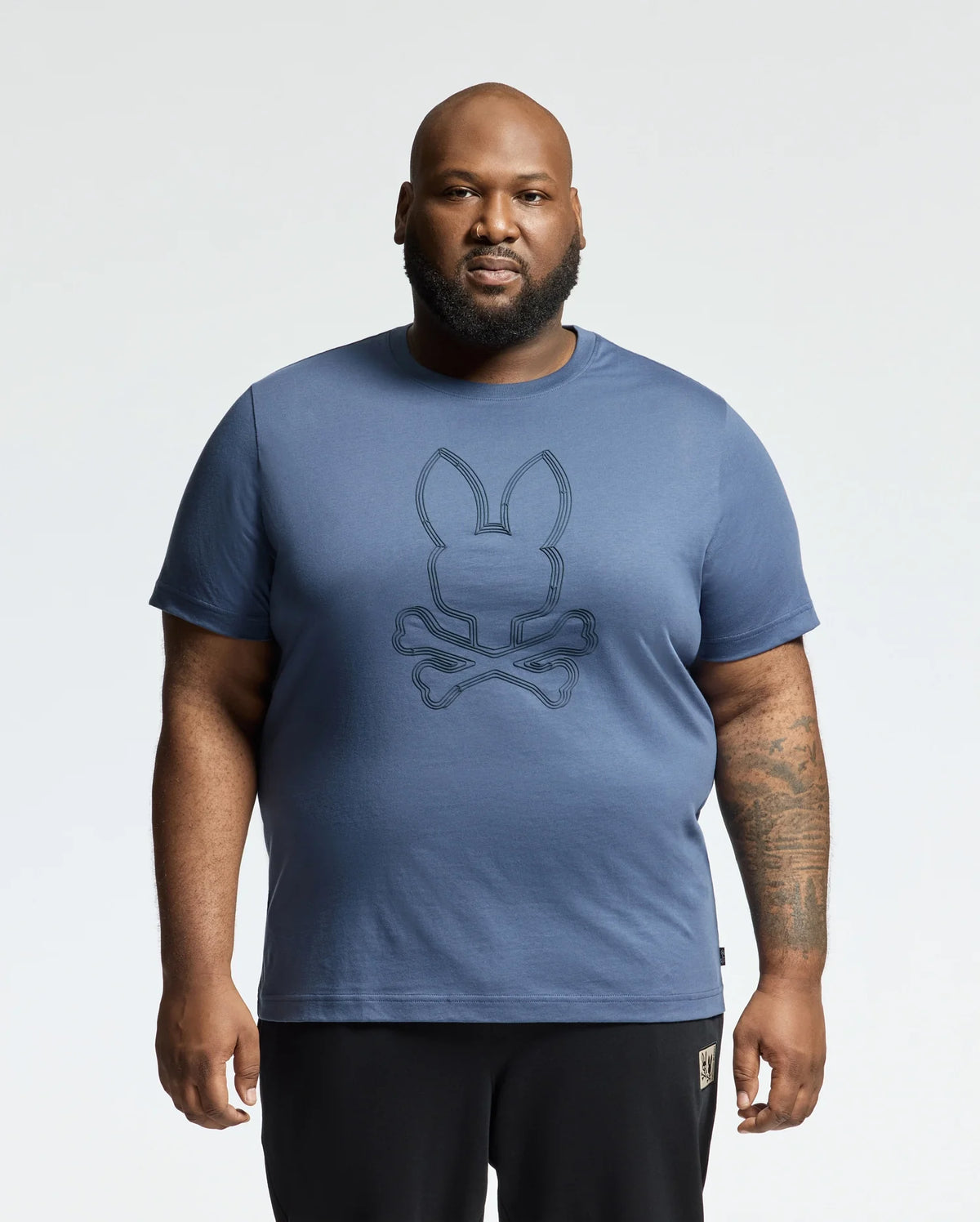 A bearded man wearing a Psycho Bunny "Mens Big and Tall Monarc Front Bunny Graphic Tee" in soft Pima cotton stands against a plain white background. The blue tee features a bunny and crossed bones design. He has a tattoo on his left forearm and pairs it with sleek black pants.