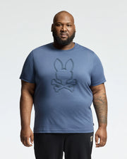 A bearded man wearing a Psycho Bunny "Mens Big and Tall Monarc Front Bunny Graphic Tee" in soft Pima cotton stands against a plain white background. The blue tee features a bunny and crossed bones design. He has a tattoo on his left forearm and pairs it with sleek black pants.