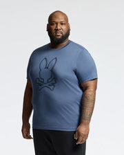 A bearded man, with tattoos on his right arm, holds a small object in his left hand while showcasing a Psycho Bunny MENS BIG AND TALL MONARC FRONT BUNNY GRAPHIC TEE - B9U211E200 made from soft Pima cotton against a plain background.