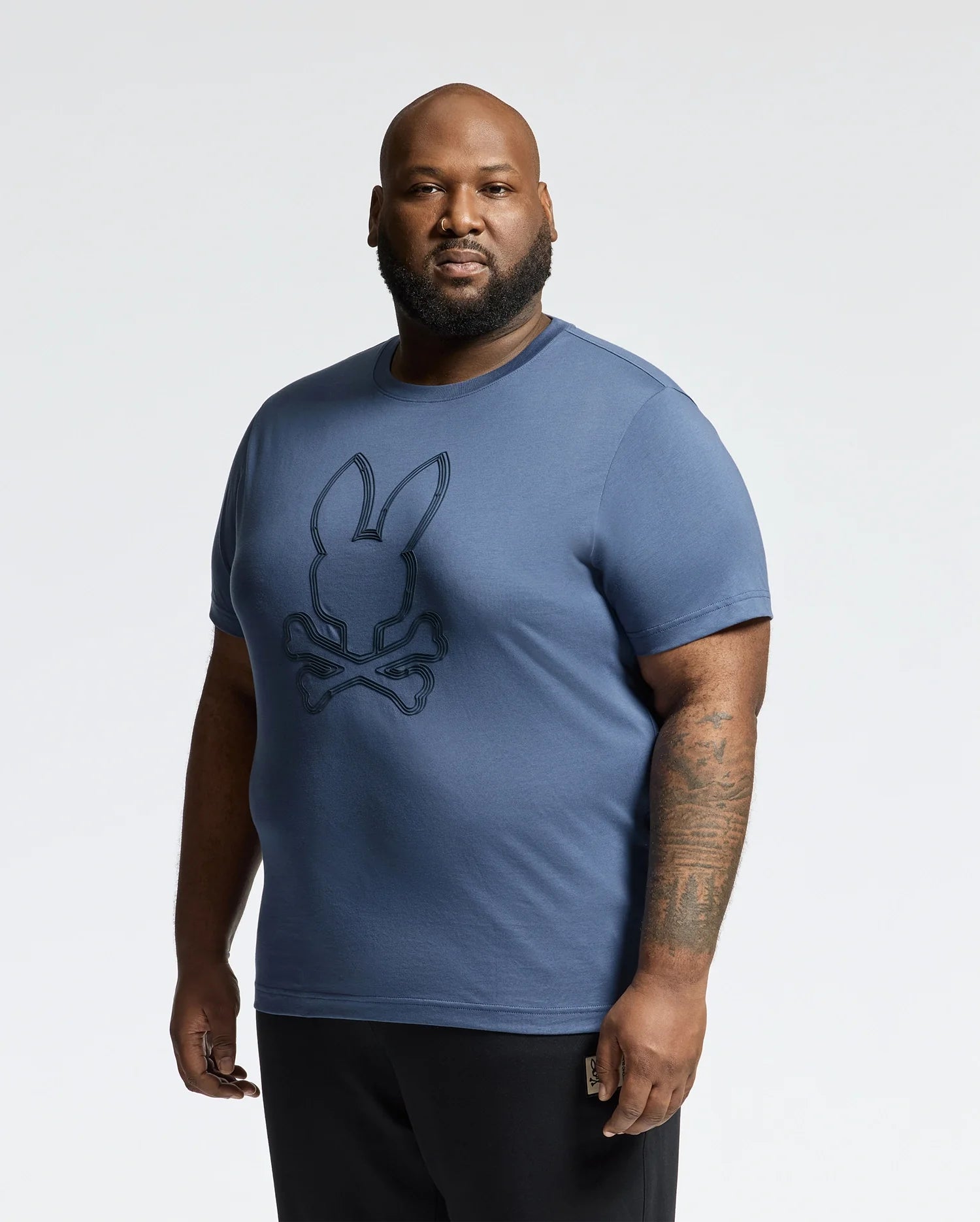 A bearded man, with tattoos on his right arm, holds a small object in his left hand while showcasing a Psycho Bunny MENS BIG AND TALL MONARC FRONT BUNNY GRAPHIC TEE - B9U211E200 made from soft Pima cotton against a plain background.