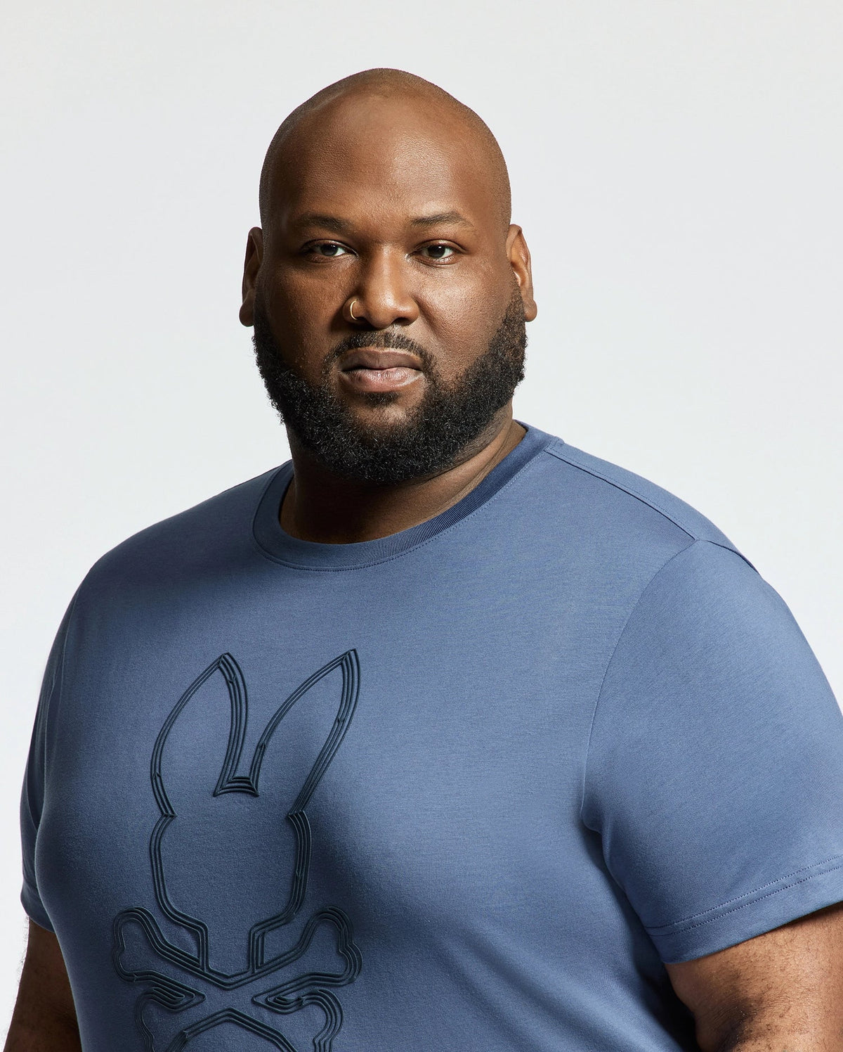 A bearded man wearing a Psycho Bunny Mens Big and Tall Monarc Front Bunny Graphic Tee - B9U211E200 in blue, featuring a stylized rabbit head with crossed bones, stands against a plain white background.