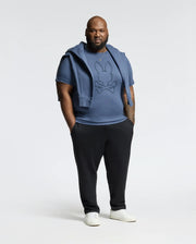 A person in a blue Psycho Bunny mens big and tall Monarc front Bunny graphic tee (B9U211E200), black pants, and white sneakers stands against a plain background. A matching blue sweater is draped over their shoulders, with hands casually tucked in pockets.