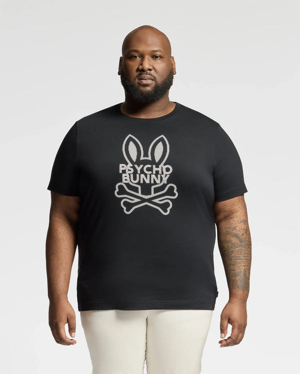 A bearded man, donning the Psycho Bunny BIG AND TALL DUMON GRAPHIC TEE (B9U271E200) with a bunny skull and crossbones design, stands against a white backdrop. This high-quality Pima Cotton tee effortlessly showcases his edgy style.