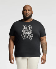 A bearded man, donning the Psycho Bunny BIG AND TALL DUMON GRAPHIC TEE (B9U271E200) with a bunny skull and crossbones design, stands against a white backdrop. This high-quality Pima Cotton tee effortlessly showcases his edgy style.
