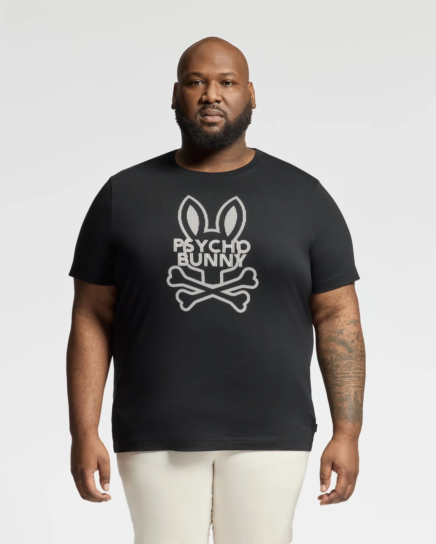 A bearded man, donning the Psycho Bunny BIG AND TALL DUMON GRAPHIC TEE (B9U271E200) with a bunny skull and crossbones design, stands against a white backdrop. This high-quality Pima Cotton tee effortlessly showcases his edgy style.