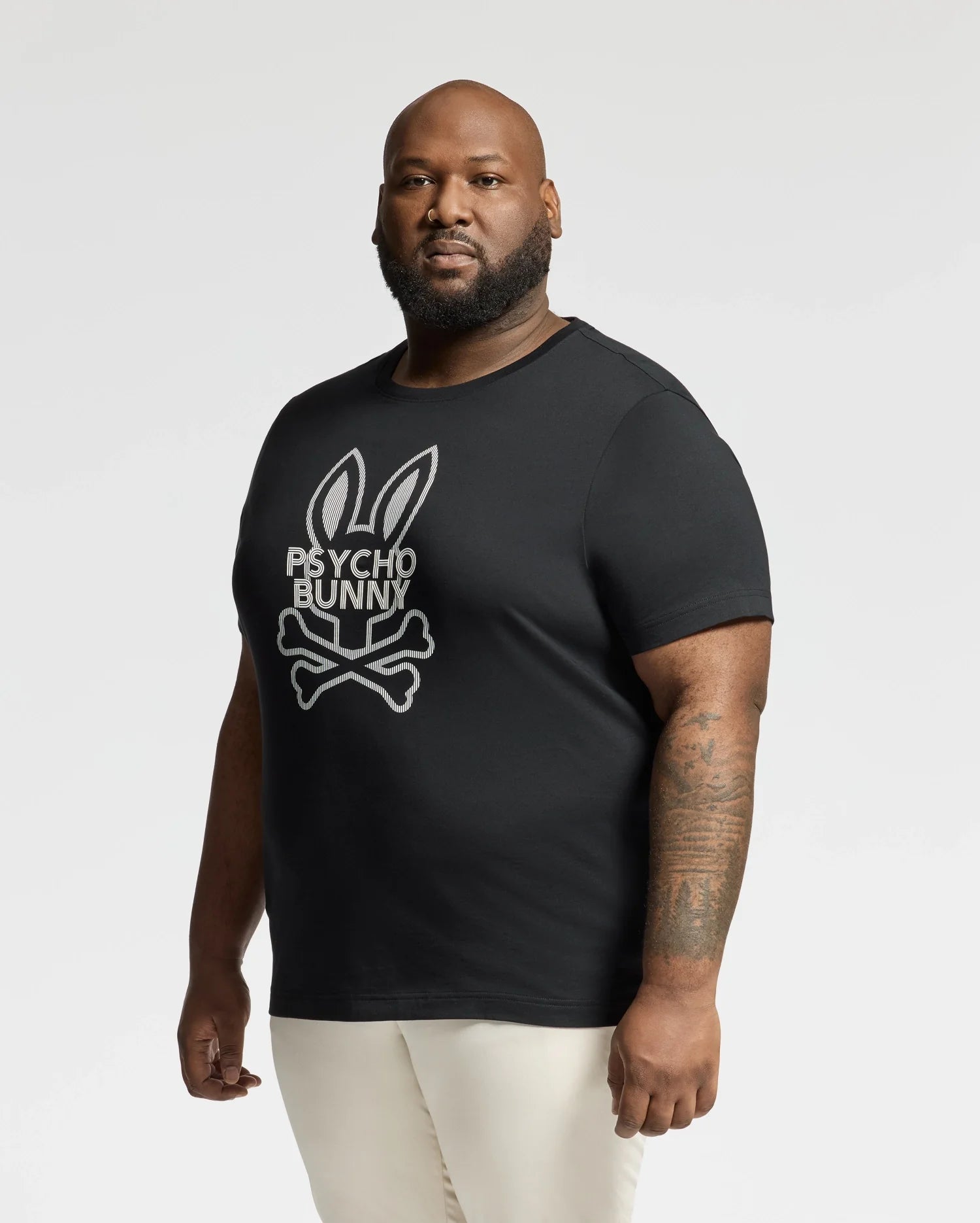 A bearded, tattooed man is wearing a premium "Psycho Bunny" BIG AND TALL DUMON GRAPHIC TEE (B9U271E200) in black, featuring a large bunny and crossbones logo. Made from soft Pima cotton, he looks directly at the camera against a plain white background.