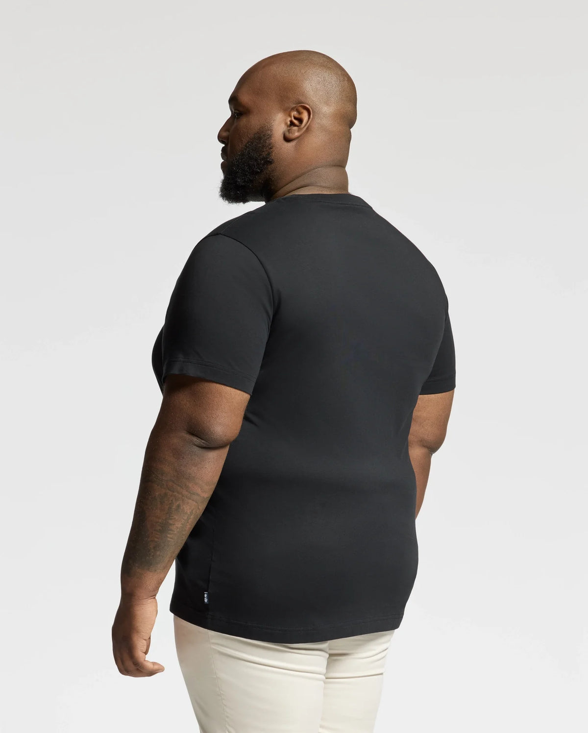 A bearded individual stands sideways, sporting the BIG AND TALL DUMON GRAPHIC TEE by Psycho Bunny in black and light-colored pants. The background is plain and light.