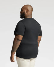 A bearded individual stands sideways, sporting the BIG AND TALL DUMON GRAPHIC TEE by Psycho Bunny in black and light-colored pants. The background is plain and light.