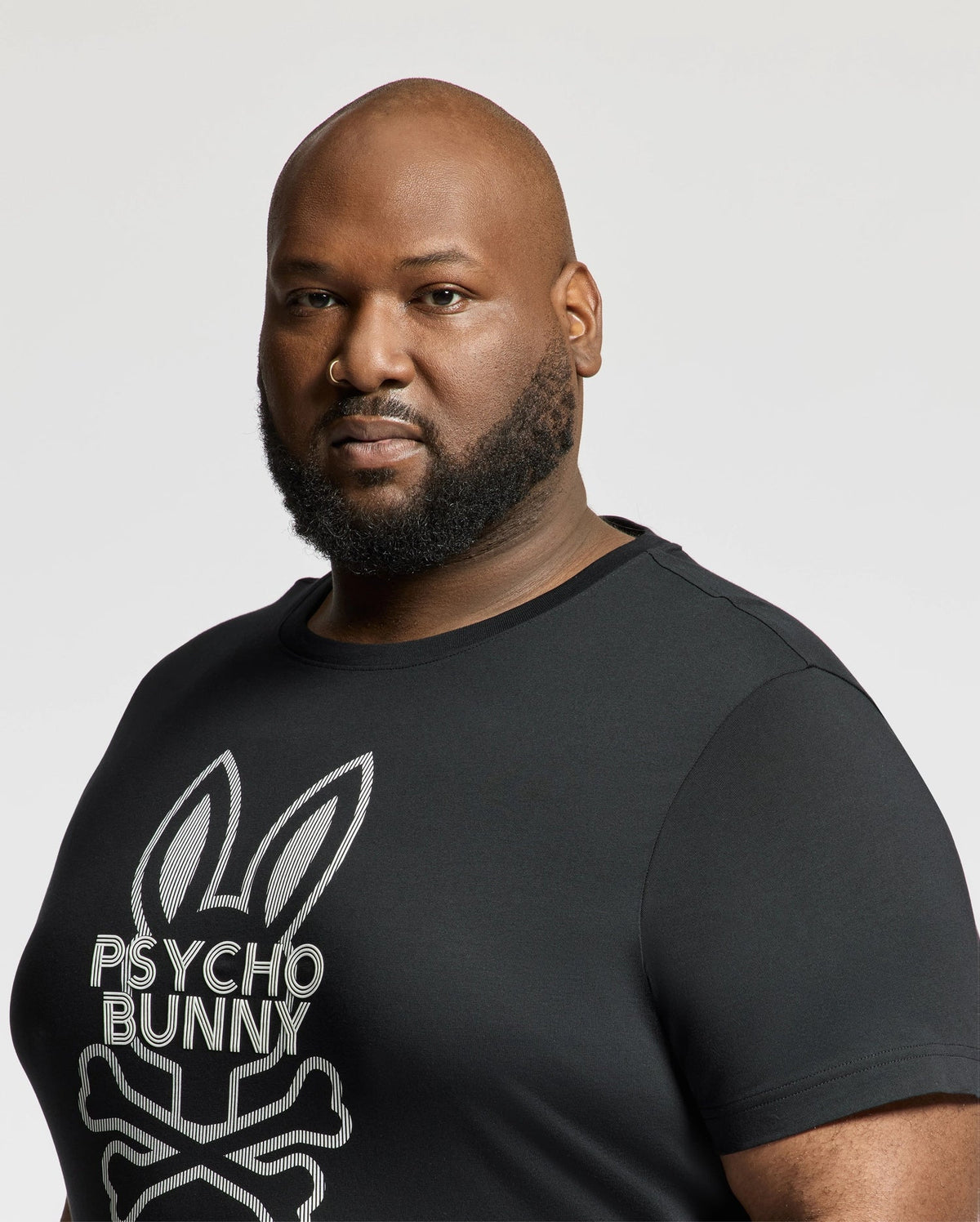 A bearded person with a shaved head wears the Psycho Bunny BIG AND TALL DUMON GRAPHIC TEE - B9U271E200, featuring a stylized bunny and crossbones design on premium Pima cotton, standing out against the plain white background.