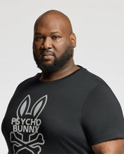 A bearded person with a shaved head wears the Psycho Bunny BIG AND TALL DUMON GRAPHIC TEE - B9U271E200, featuring a stylized bunny and crossbones design on premium Pima cotton, standing out against the plain white background.