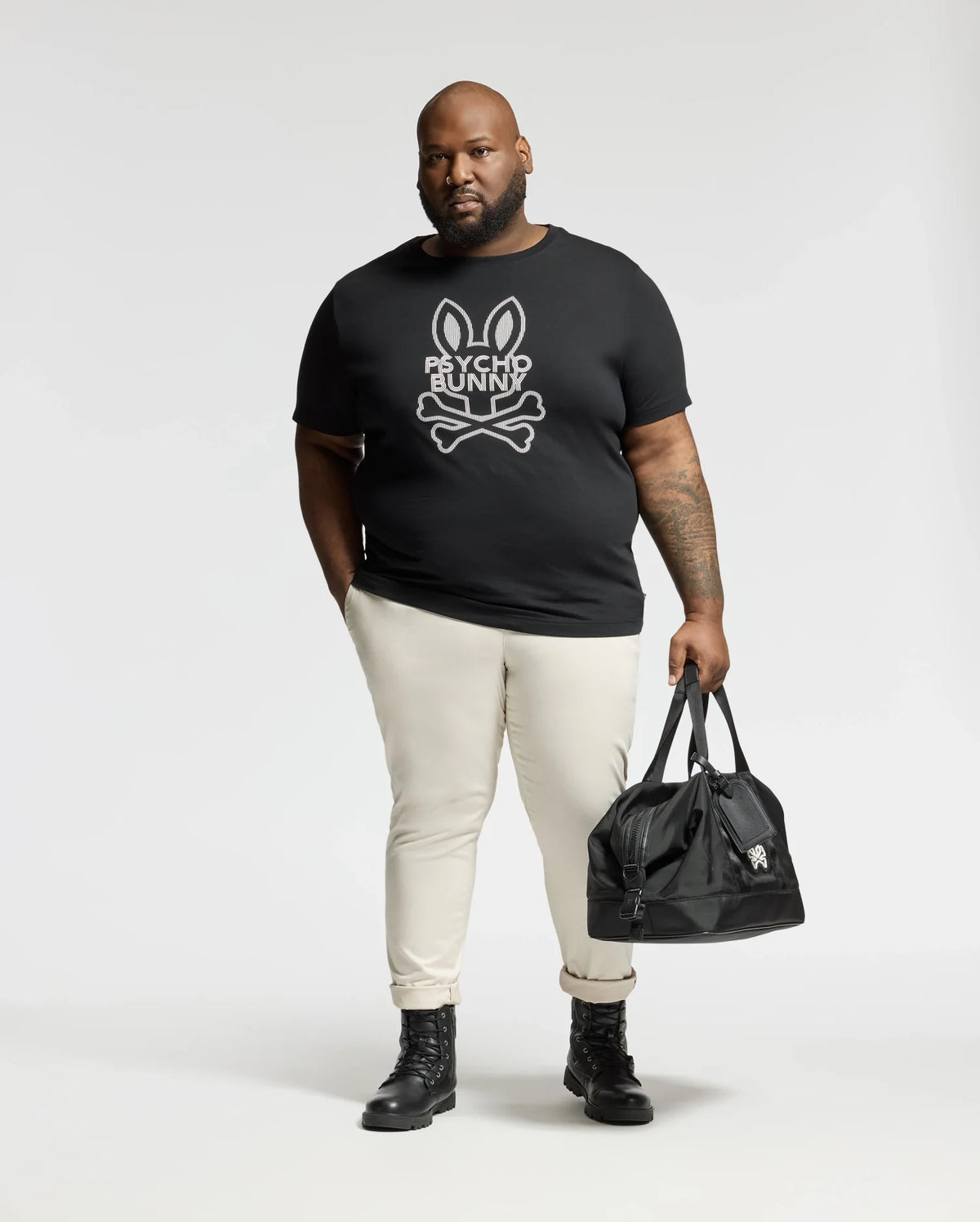 A man stands against a plain background wearing the Psycho Bunny BIG AND TALL DUMON GRAPHIC TEE - B9U271E200 paired with light-colored pants and black boots, carrying a sleek black duffel bag.