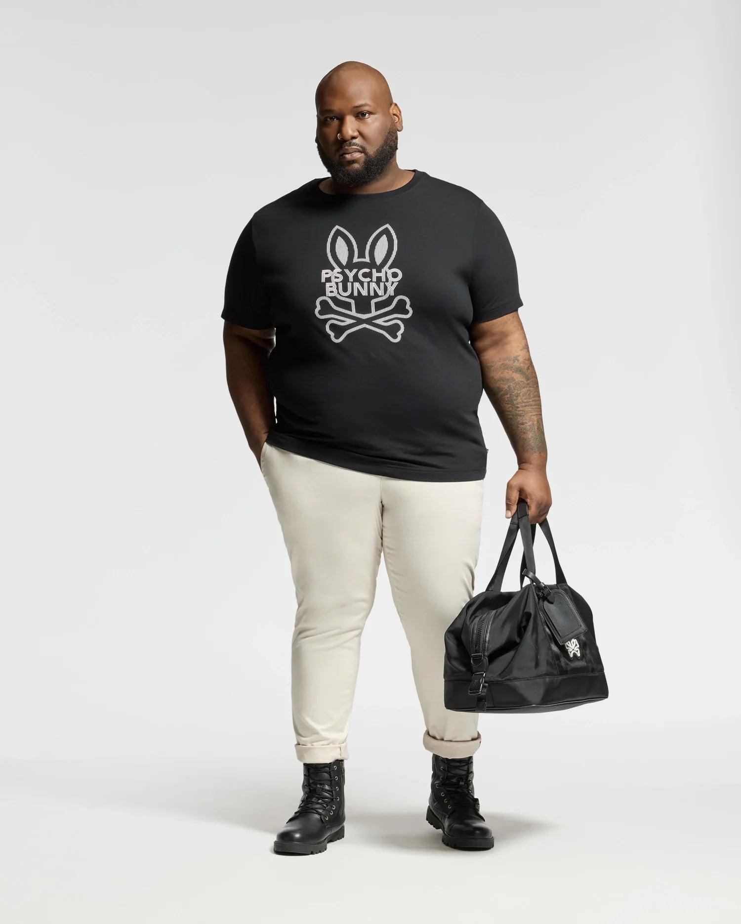 A man stands against a plain background wearing the Psycho Bunny BIG AND TALL DUMON GRAPHIC TEE - B9U271E200 paired with light-colored pants and black boots, carrying a sleek black duffel bag.