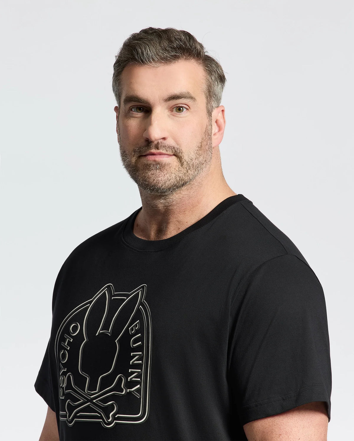 A bearded man with short hair shows off a serious look in the eco-friendly black Psycho Bunny Men's Big and Tall Melbourne Graphic Tee, featuring a bunny and crossbones design. The premium feel of its plain white background is crafted from Peruvian Pima cotton for ultimate comfort.