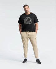 A man wears a black Psycho Bunny Big and Tall Melbourne Graphic Tee (B9U410F200), made from eco-friendly Peruvian Pima cotton, along with beige cargo pants. Standing against a plain background, he has his hands in his pockets and a neutral expression.