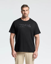 A man with short hair and a beard wears the Psycho Bunny MENS BIG AND TALL BERNI EMBROIDERED WORDMARK GRAPHIC TEE on Pima cotton, featuring an embroidered graphic. He pairs it with beige pants against a plain light background.