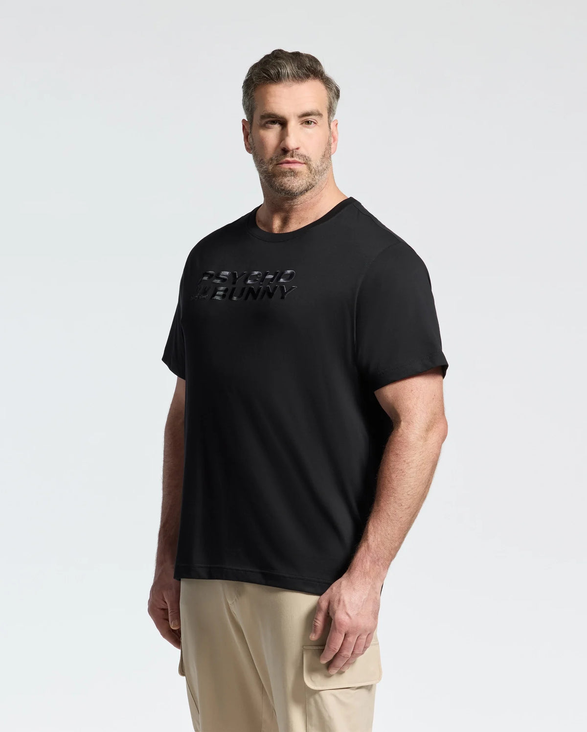 A bearded man with short hair wearing the Psycho Bunny MENS BIG AND TALL BERNI EMBROIDERED WORDMARK GRAPHIC TEE - B9U417F200 and beige cargo pants stands against a plain light background, the raised text on his black Pima cotton tee visible as he looks directly at the camera.
