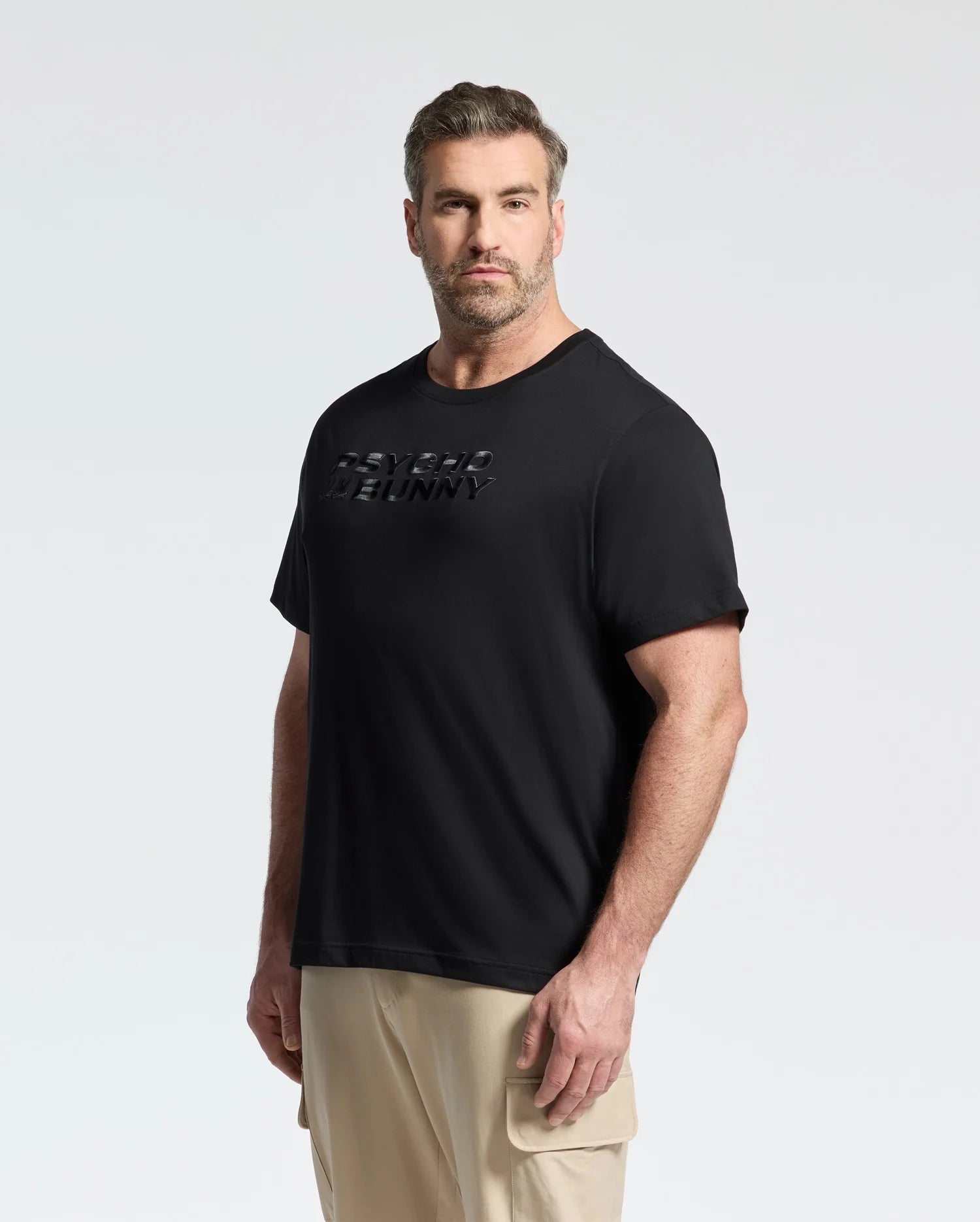 A bearded man with short hair wearing the Psycho Bunny MENS BIG AND TALL BERNI EMBROIDERED WORDMARK GRAPHIC TEE - B9U417F200 and beige cargo pants stands against a plain light background, the raised text on his black Pima cotton tee visible as he looks directly at the camera.