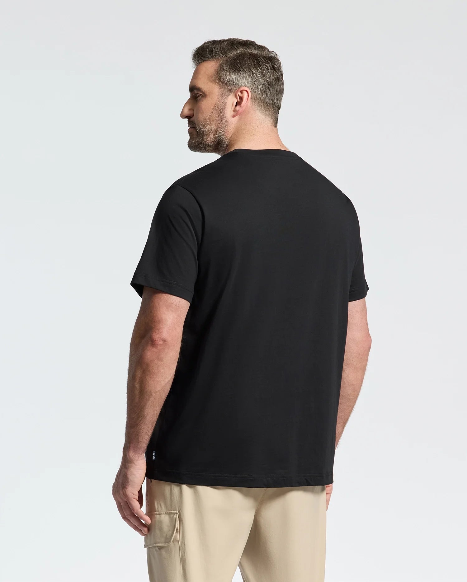A short-haired man faces away, wearing a breathable Psycho Bunny black tee and beige cargo shorts, set against a plain light gray background.