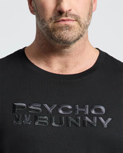 A man is wearing the Psycho Bunny Men's Big and Tall Berni Embroidered Wordmark Graphic Tee with bold lettering and small embroidered bunny logos. Only his lower face and upper body are visible.
