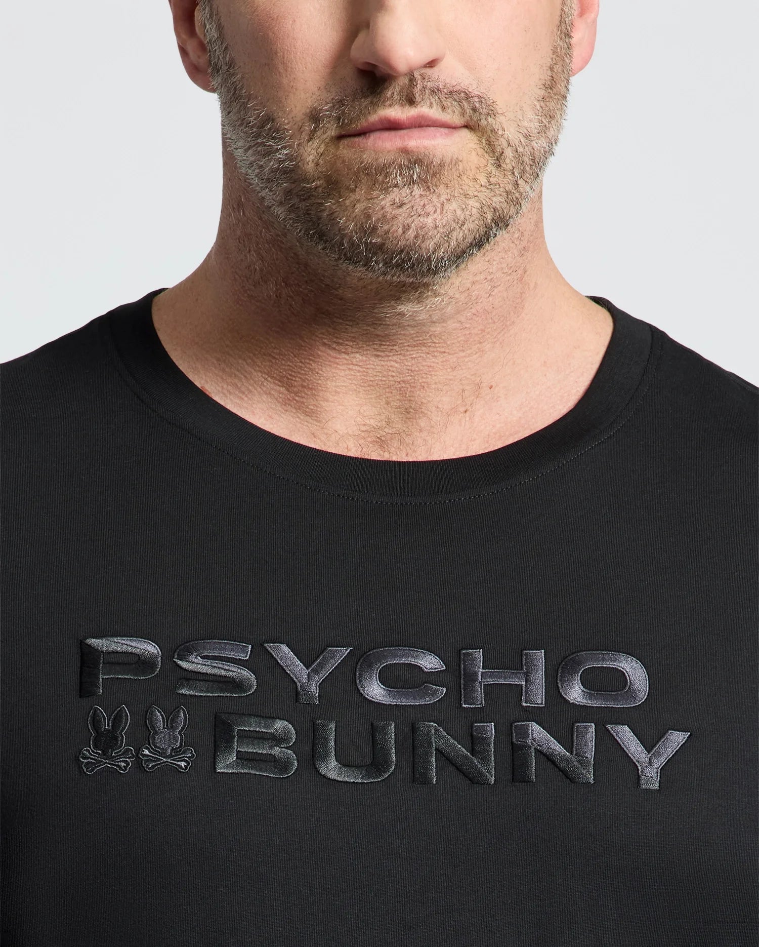 A man is wearing the Psycho Bunny Men's Big and Tall Berni Embroidered Wordmark Graphic Tee with bold lettering and small embroidered bunny logos. Only his lower face and upper body are visible.