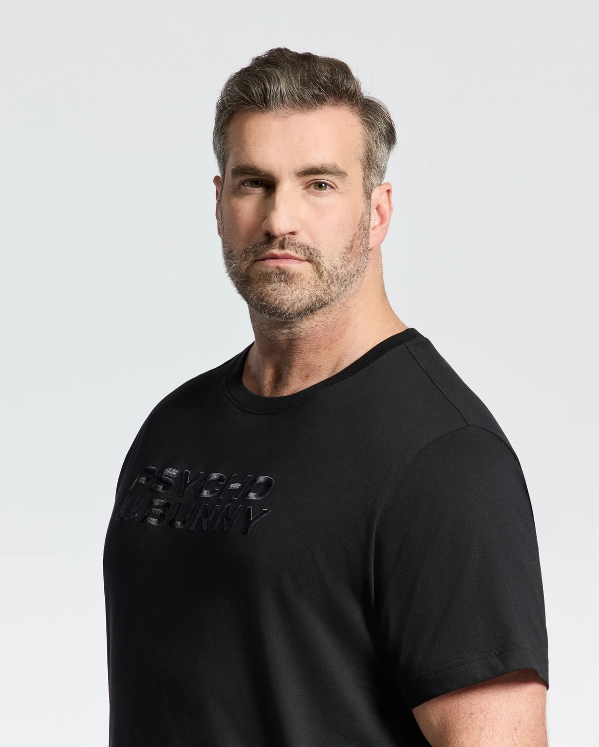 A bearded man with short gray hair dons the Psycho Bunny MENS BIG AND TALL BERNI EMBROIDERED WORDMARK GRAPHIC TEE, B9U417F200. The black Pima cotton tee features textured lettering as he gazes at the camera against a plain white background, exuding effortless style.