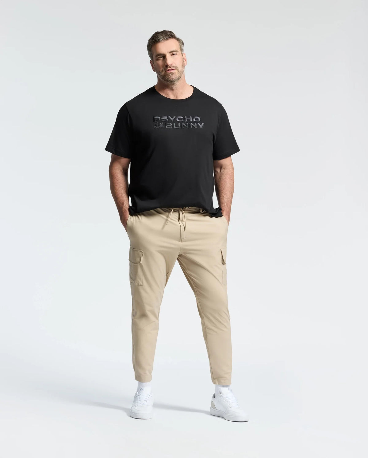 A man with short hair confidently stands wearing a Psycho Bunny MENS BIG AND TALL BERNI EMBROIDERED WORDMARK GRAPHIC TEE in black Pima cotton, paired with beige cargo pants. His hands are in his pockets, and he sports white sneakers against a plain white background.