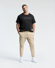 A man with short hair confidently stands wearing a Psycho Bunny MENS BIG AND TALL BERNI EMBROIDERED WORDMARK GRAPHIC TEE in black Pima cotton, paired with beige cargo pants. His hands are in his pockets, and he sports white sneakers against a plain white background.
