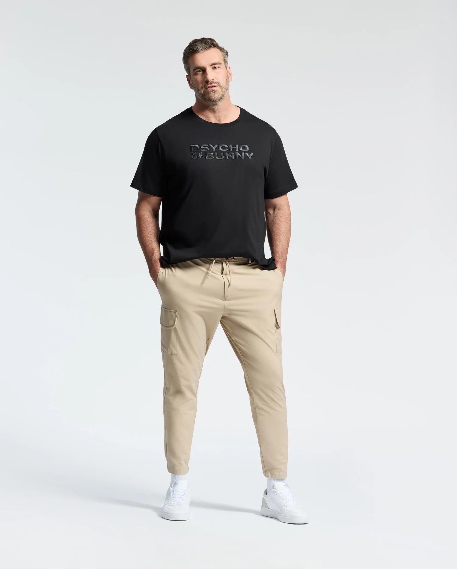 A man with short hair confidently stands wearing a Psycho Bunny MENS BIG AND TALL BERNI EMBROIDERED WORDMARK GRAPHIC TEE in black Pima cotton, paired with beige cargo pants. His hands are in his pockets, and he sports white sneakers against a plain white background.