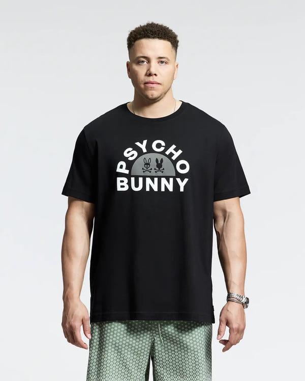 Wearing a sleek black MENS BIG AND TALL GOLDIE GRAPHIC TEE - B9U606F200 from Psycho Bunny, a person stands against a plain backdrop. The tee features "Psycho Bunny" and two small bunnies. They pair it with patterned shorts and a wristwatch for an effortlessly stylish look.
