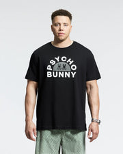 Wearing a sleek black MENS BIG AND TALL GOLDIE GRAPHIC TEE - B9U606F200 from Psycho Bunny, a person stands against a plain backdrop. The tee features "Psycho Bunny" and two small bunnies. They pair it with patterned shorts and a wristwatch for an effortlessly stylish look.