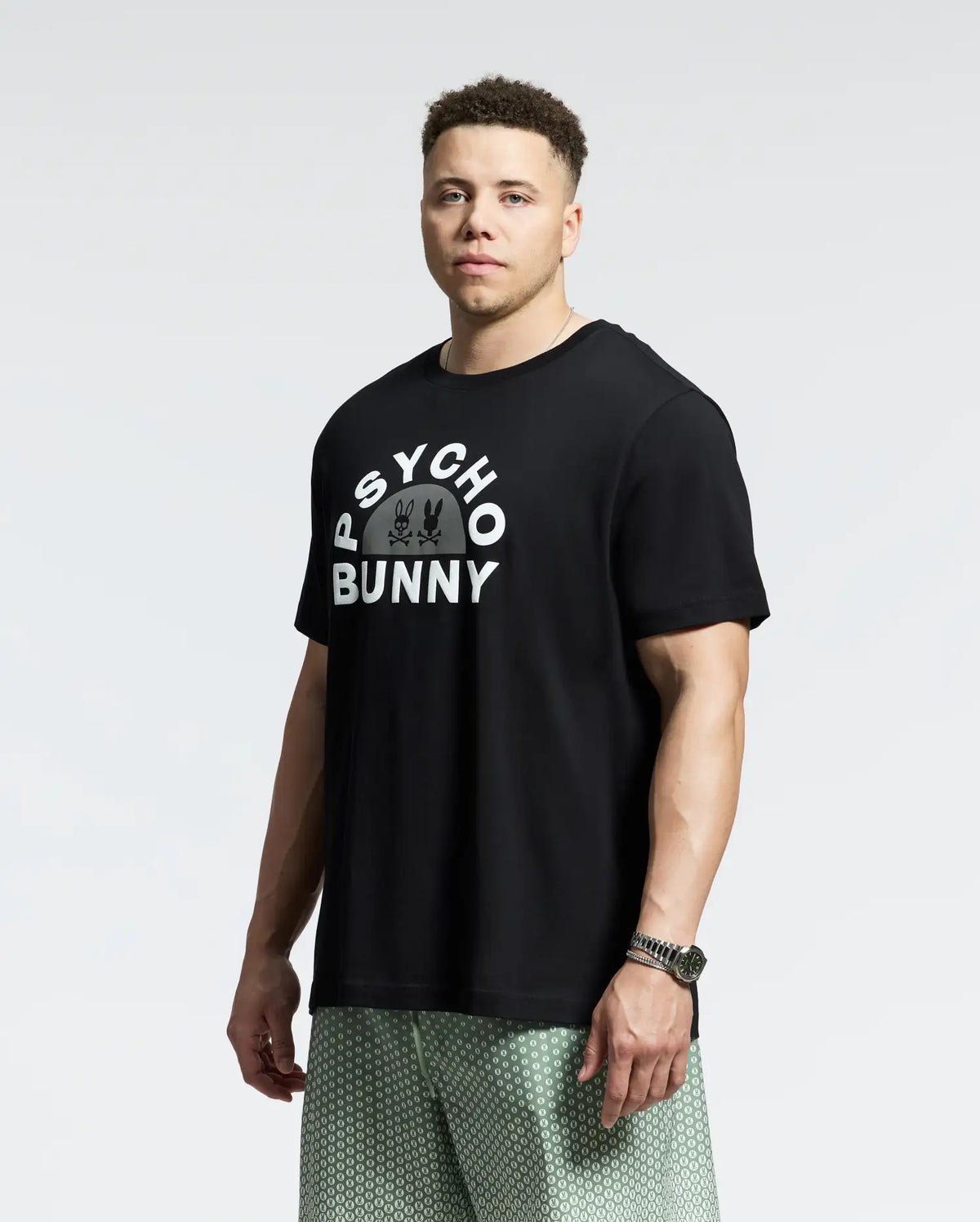 A man with short hair stands against a white background wearing the MENS BIG AND TALL GOLDIE GRAPHIC TEE - B9U606F200 by Psycho Bunny, crafted from soft Pima cotton, featuring the iconic Bunny graphic in white letters. He pairs it with patterned green shorts and a watch.