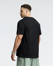 A man with short curly hair stands, glancing over his shoulder in a black Psycho Bunny Goldie Graphic Tee and patterned green shorts. A watch adorns his wrist, and the minimalistic white backdrop accentuates his cool style.